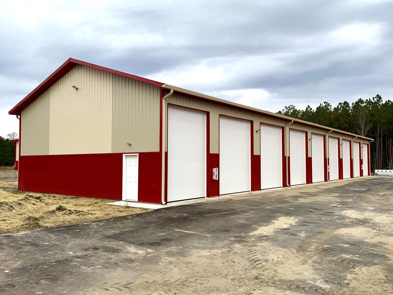 A beige and red metal storage building with nine white garage doors, a small white door, and a dirt ground